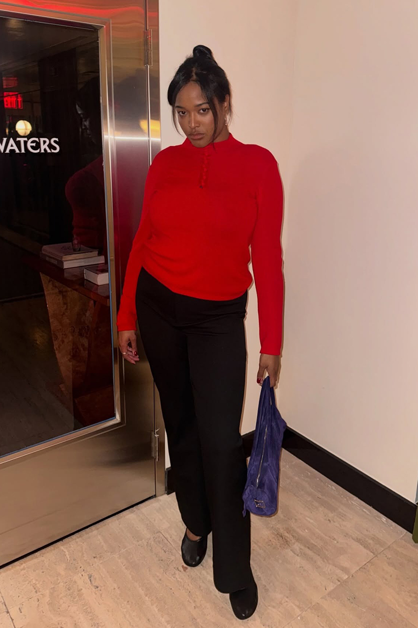 a woman wearing a red longsleeve polo, black pants, black flats, and a purple barrel suede bag
