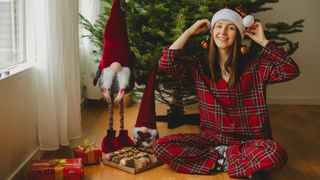 A young woman wearing tartan Christmas pajamas and a Santa hat decorates a Christmas tree. 