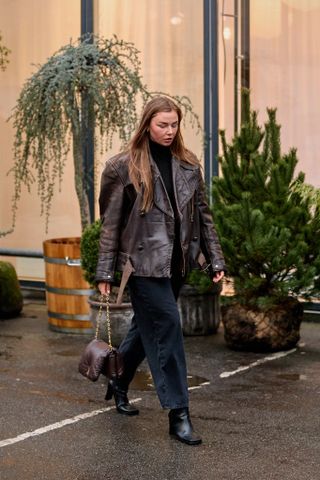 how to wear cropped flare jeans shown on a woman walking wearing black jeans with a black turtleneck, brown leather jacket, and brown leather boots