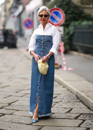 Grece Ghanem wears black vintage sunglasses, white pearl pendant earrings, a blue denim shoulder-off / buttoned long ripped dress, a pale yellow shiny leather braided Jodie handbag from Bottega Veneta, blue satin/silk pointed pumps heels shoes, and a silver large bracelet outside Sportmax on February 24, 2023, during the Milan Fashion Week Womenswear Fall/Winter 2023/2024 in Milan, Italy.