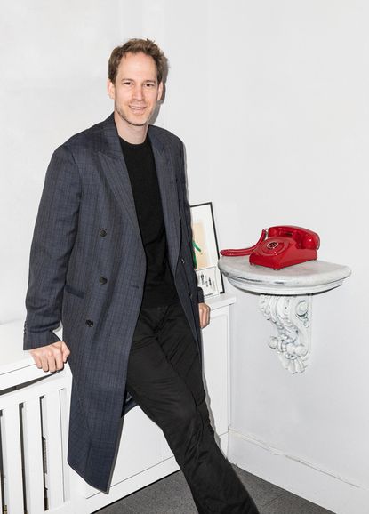 A close-up of David Korins wearing a black jumper and grey blazer standing next to an old red corded telephone. 