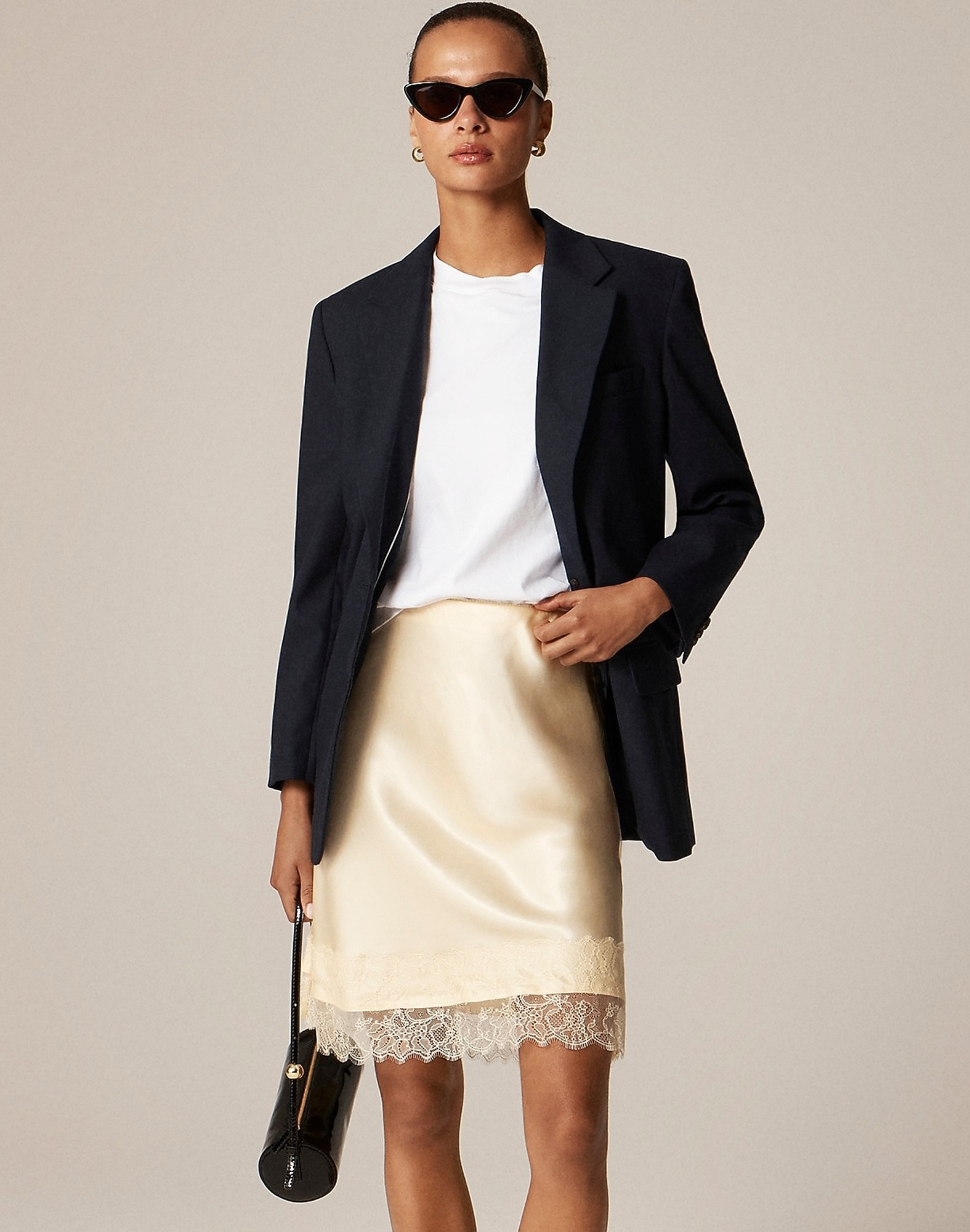 J.Crew fall 2025 outfit idea with black cat-=eye sunglasses, a black blazer, a white T-shirt, pale yellow satin skirt with lace hem, and small black duffle bag