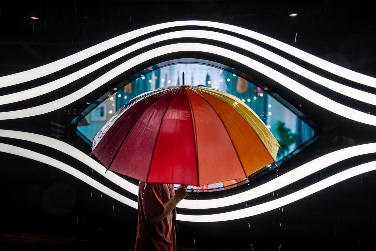 Umbrella Eye - A person walks past a giant eye on a shopping facade, with a colorful umbrella positioned perfectly at the center, creating the illusion of an &#039;umbrella eye.&#039; 