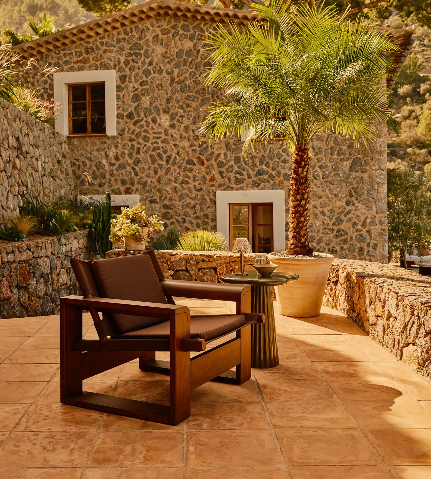 IMage of a large brown outdoor accent chair on a stucco patio with a green lacquer side table beside it and a stone house in the background.