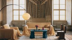 Image of a large living room with high ceilings and two large windows. There is a sofa with a pale yellow cover and draping arms, and a matching accent chair. There is a blue coffee table as well.