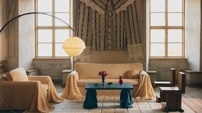 Image of a large living room with high ceilings and two large windows. There is a sofa with a pale yellow cover and draping arms, and a matching accent chair. There is a blue coffee table as well.