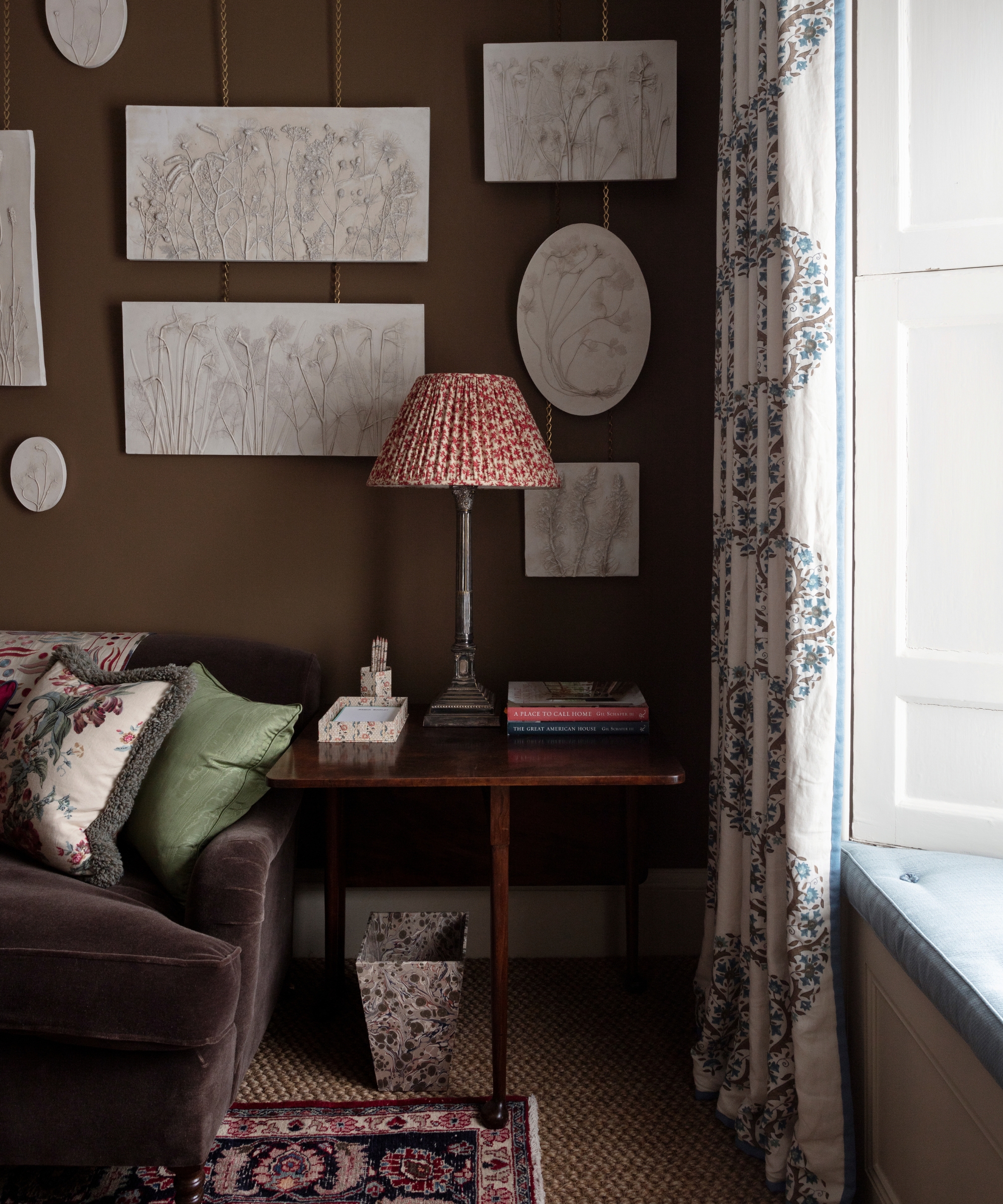 A cozy study with dark brown walls, a red floral lampshade, botanical plaster artworks and blue patterned curtains.