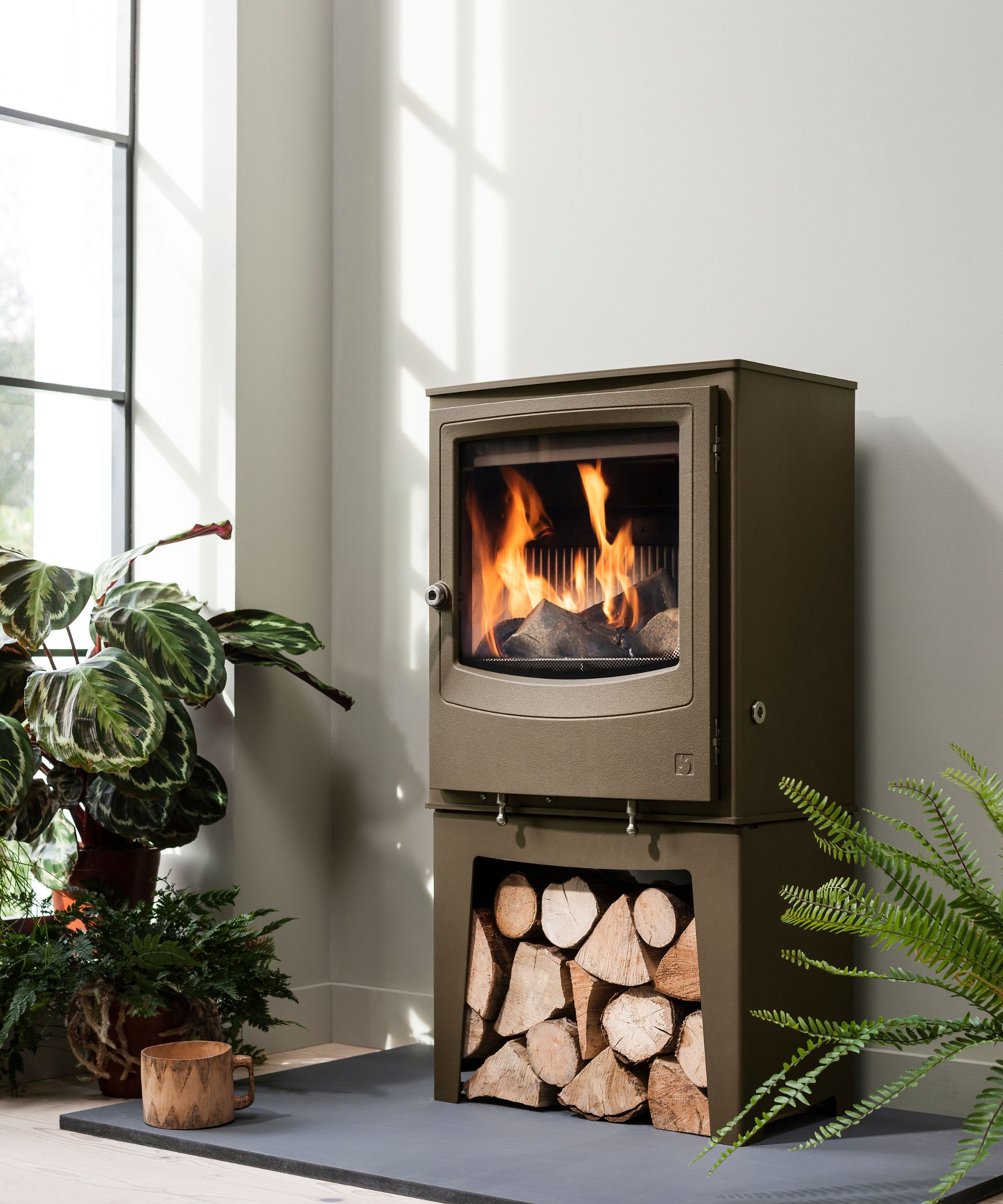 A log burner in an olive green finish stands on a dark slate hearth, with a built-in log store filled with cut wood.