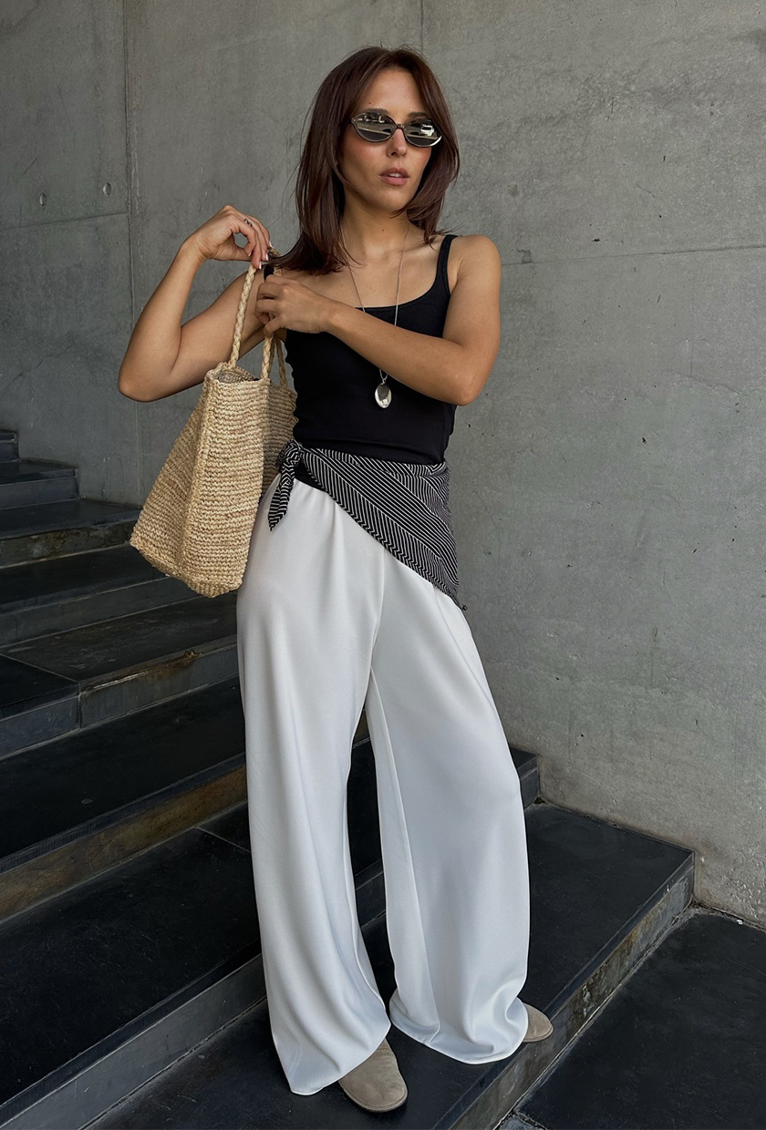 The summer aesthetic, coastal boho, is shown in a street style photo of a woman standing on a staircase wearing a black tank top with white wide leg pants, tan mesh ballet flats, a black printed scarf tied around the waist, a long silver pendant necklace, black sunglasses, and a large raffia tote bag