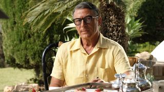 Jason Isaacs in a yellow shirt as Cary Grant sits at a table with food in front of him and a palm tree behind him in Archie.