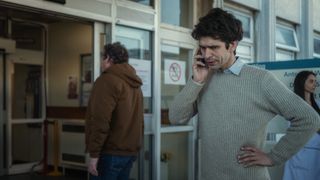 Adam (Ben Whishaw) stands outside the entrance of the hospital wearing his civilian clothes of a grey sweater over a blue shirt. He is in the middle of a call on his mobile phone and looks slightly irritated - out of focus, in the background, people are walking into and out of the hospital