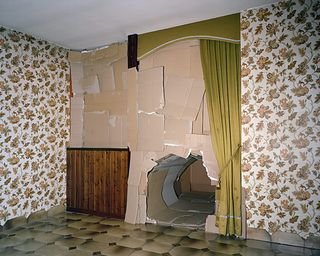 ’Mr. Schuhlmann or the Man in the High Castle’ by Matthieu Lavanchy, 2009. A wall with floral wallpaper and a large hole at the bottom of it.