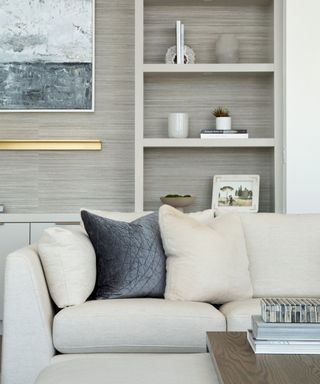 A living room with grey wallpapered wall, taupe bookshelf, and white sofa with pillows