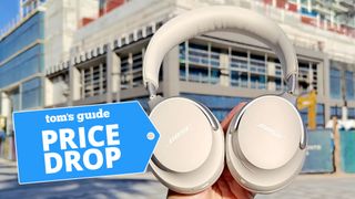 Bose QuietComfort Ultra Headphones being held up in front of urban building with price drop badge