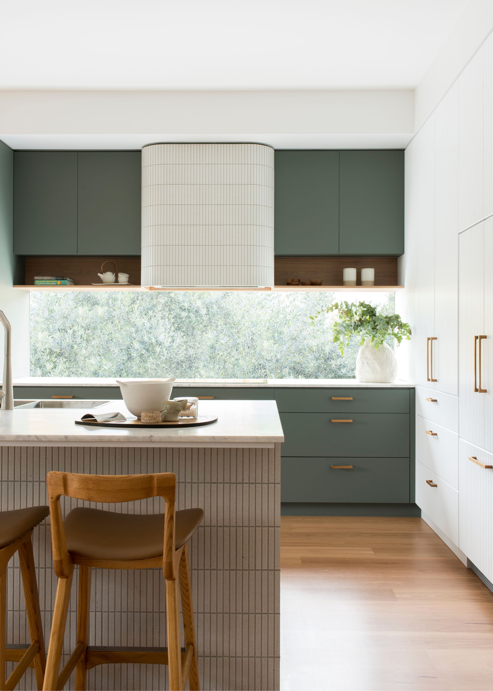 A sage green kitchen with upper and lower cabinets. There is also a kitchen island in the space with bar stools beside it.