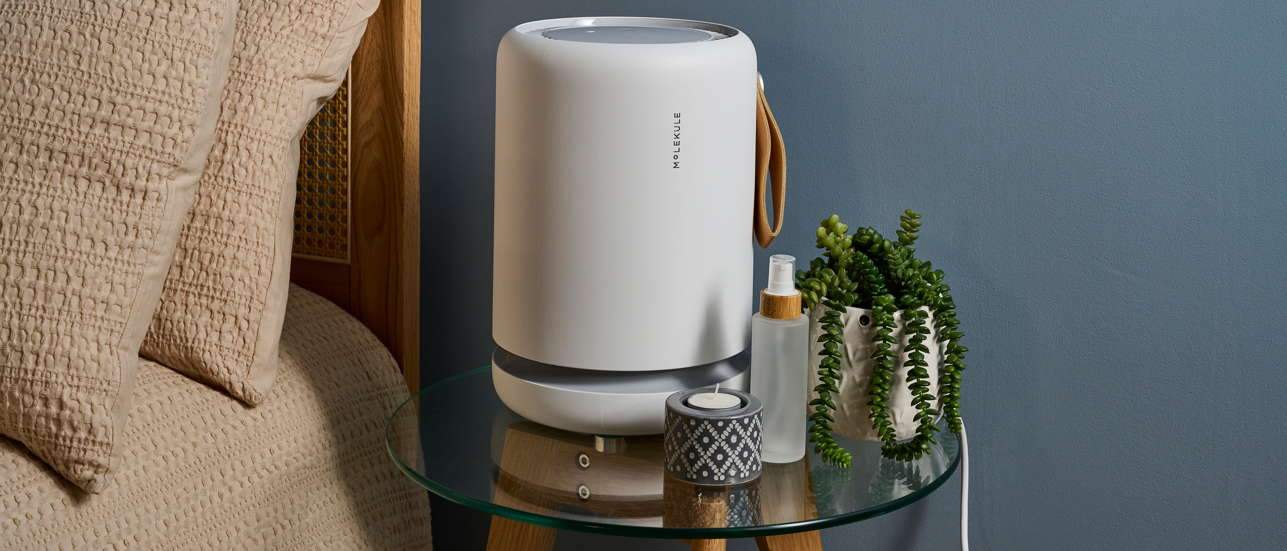 Picture shows a Molekule Air Mini Plus air purifier sitting on a glass bedside table with a candle, cosmetic bottle, and potted plant to its right.