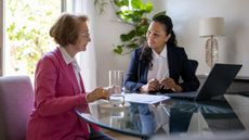 An older woman and younger woman in a suit sit at a table and discuss the paperwork that sits before them.