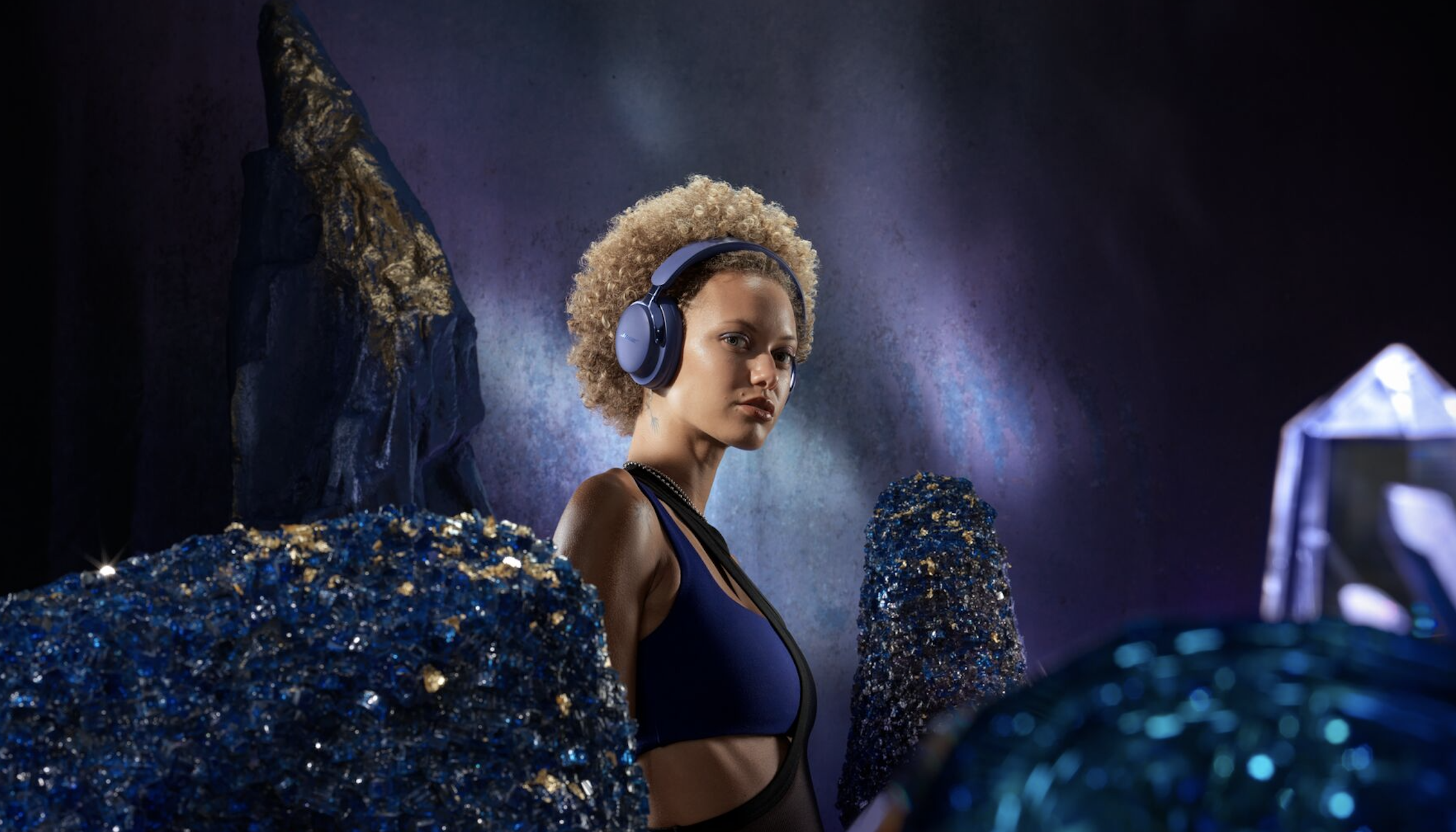 Bose QCU Headphones (2nd Gen) in &#039;Midnight Violet&#039; worn by a woman, surrounded by purple crystals