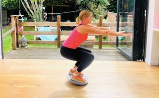 A woman is squatting on a balance board with her knees bent and arms extended in front of her. She has blonde hair in a ponytail, a pink tank top, black leggings, and orange sneakers.