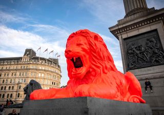 Side view of Please Feed The Lions by Es Devlin at Trafalgar Square during the day - a red lion statue on a platform with people and buildings in the background