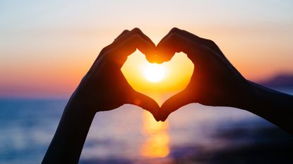 At the beach pair of hands silhouetted against the sunset forms a heart shape capturing the setting sun inside them.