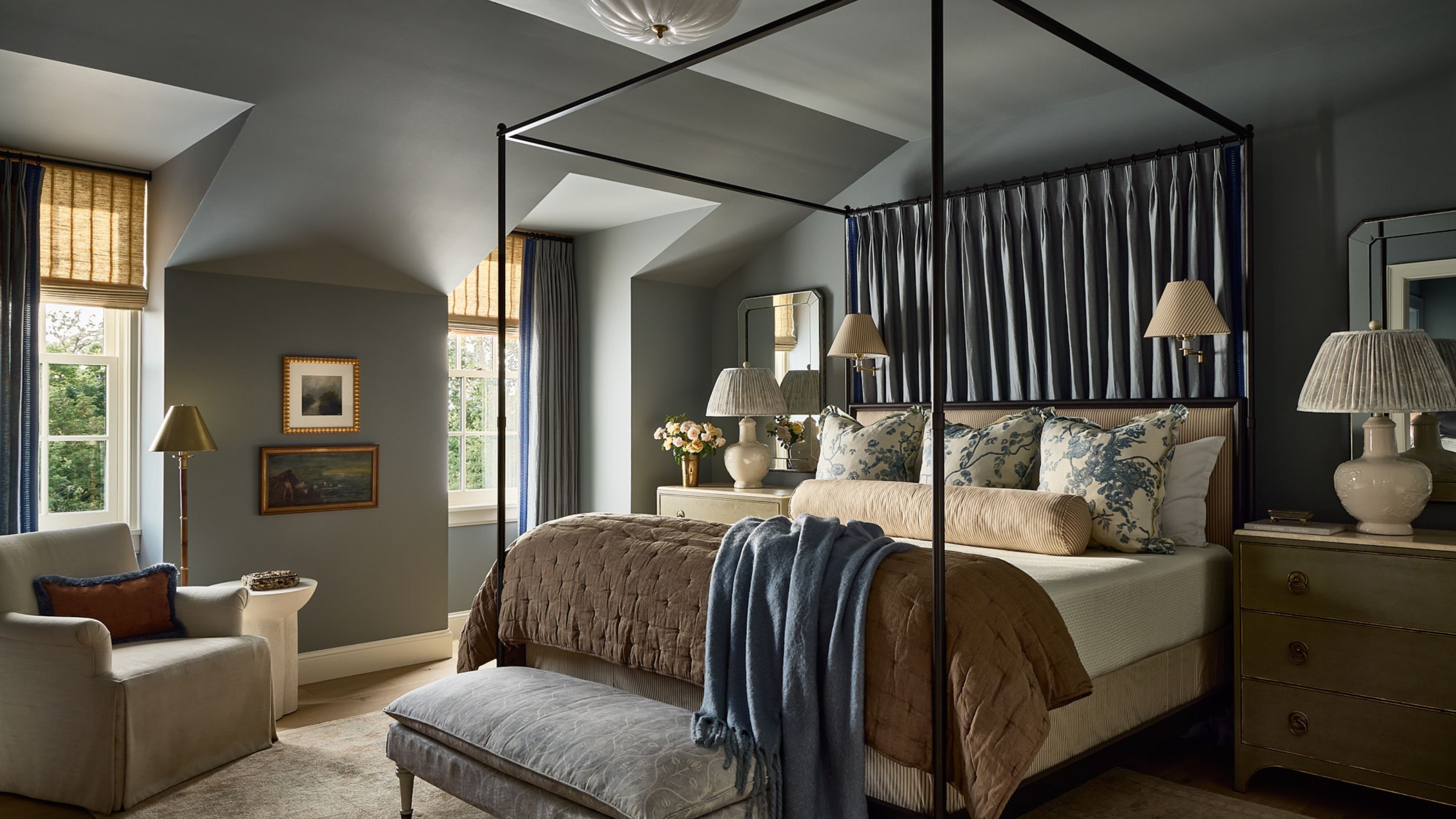 A large bedroom with blue-gray walls and ceiling, a four-poster bed with fabric draped above the headboard. A beige rug, woven window blinds, and blue curtains, a gray armchair, and floral blue cushions on the bed.