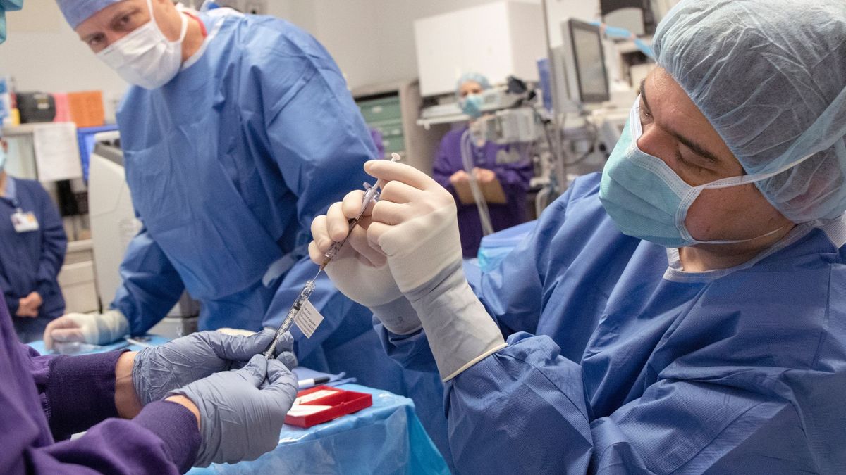 A photo shows two male doctors in surgery garb as one preps a long needle for a procedure. A patient is under a blue sheet on an operating table but can&#039;t be seen.