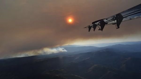 The wing of an airplane hangs over the right side of the image, a dimmed sun shines behind brown smoke above a dark, mountainous landscape.