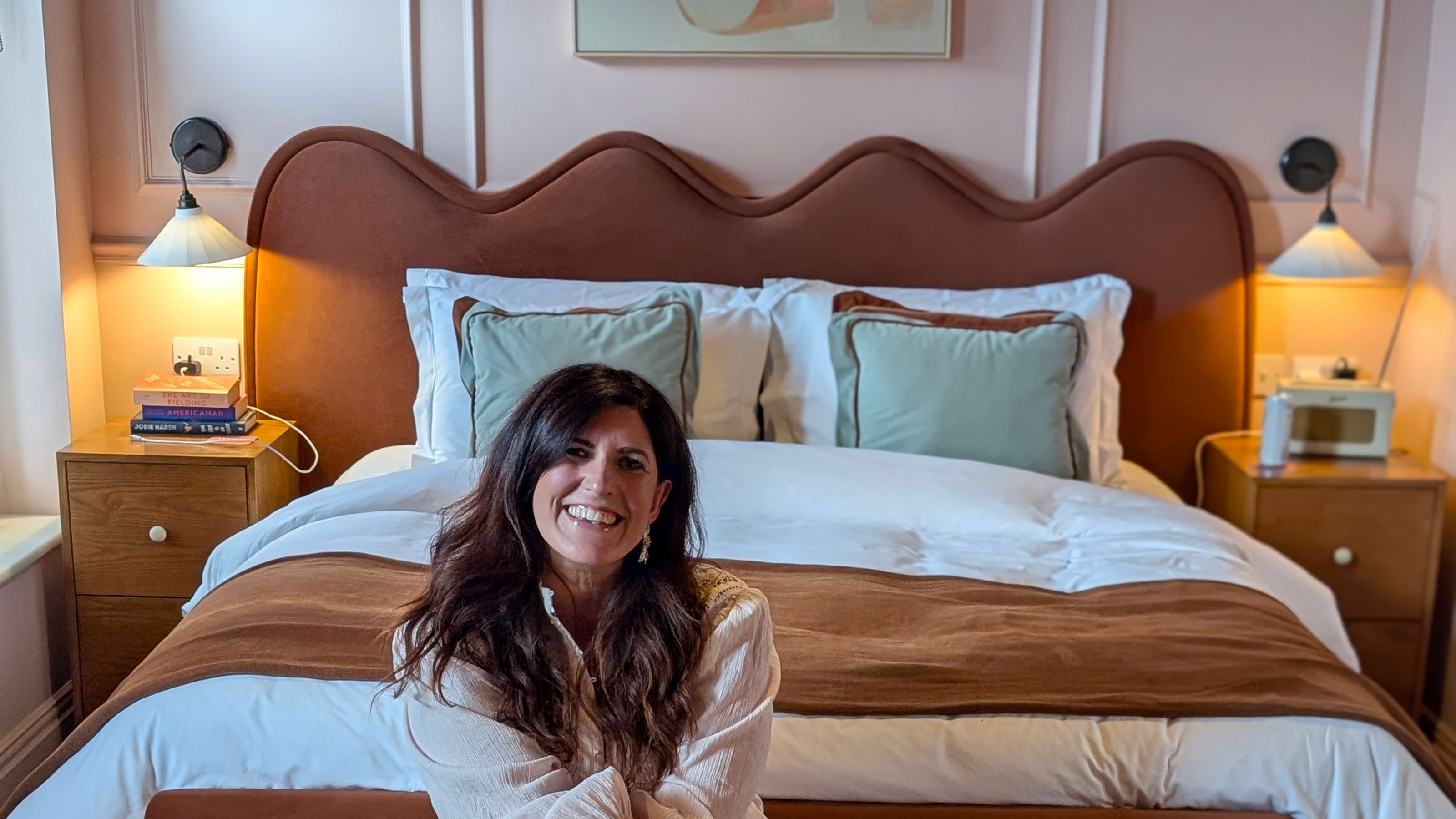 Woman sitting on edge of made bed with brown, wavy headboard in pink panelled room