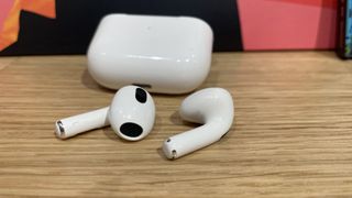 A pair of Apple AirPods 3 lying in front of their case on a wooden surface.