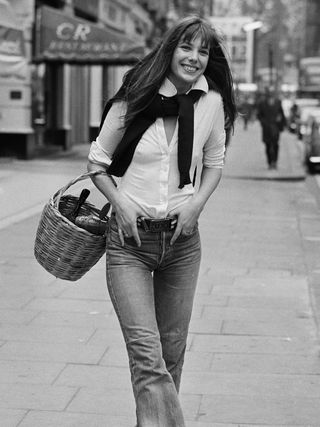 Jane Birkin wearing jeans and a white button down carrying a basket in a black and white photograph