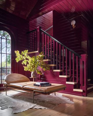Entryway with glossy burgundy painted panelled walls and staircase, tan leather daybed and green foliage in clay pot