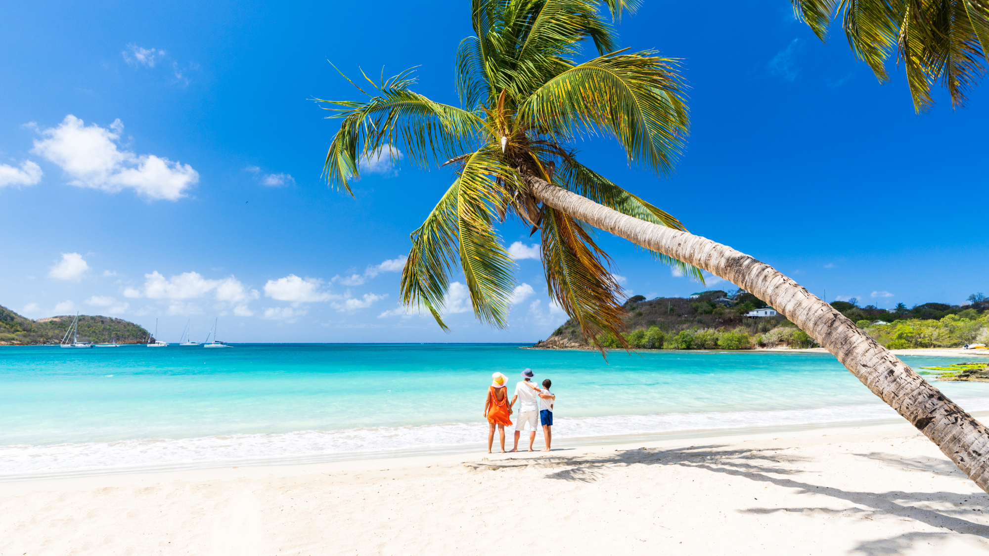 Woman and two children stand on white sandy beach in Antigua with turquoise sea and palm trees