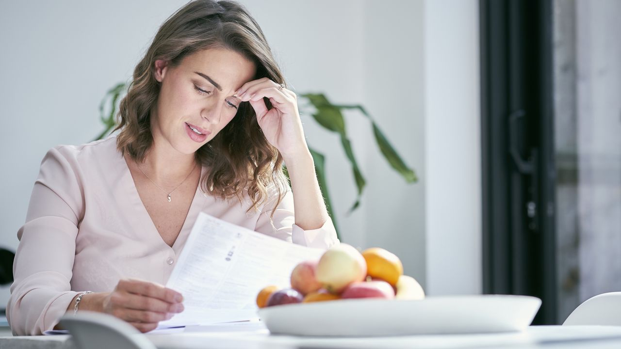 A woman looks stressed as she looks at paperwork while sitting at her dining room table.