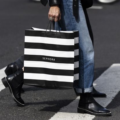 A person photographed from the waist down wearing blue jeans and black boots carrying a black and white striped Sephora shopping bag