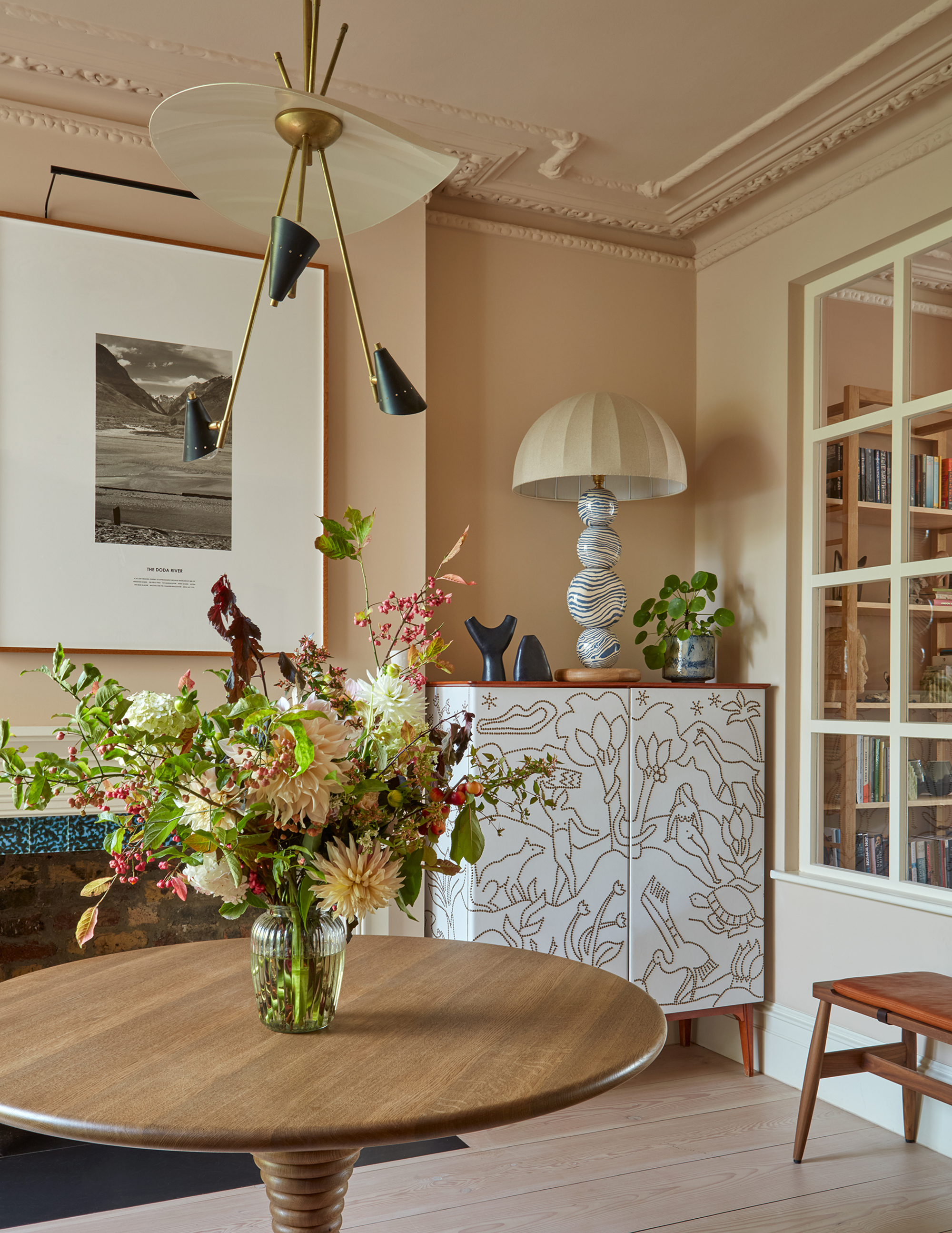 neutral living space with round timber table with a vase of flowers, white cabinet with drawn-on motifs, styled with a lamp, potted plant, and sculptures, neutral walls, large artwork over fireplace, and sculptural pendant light