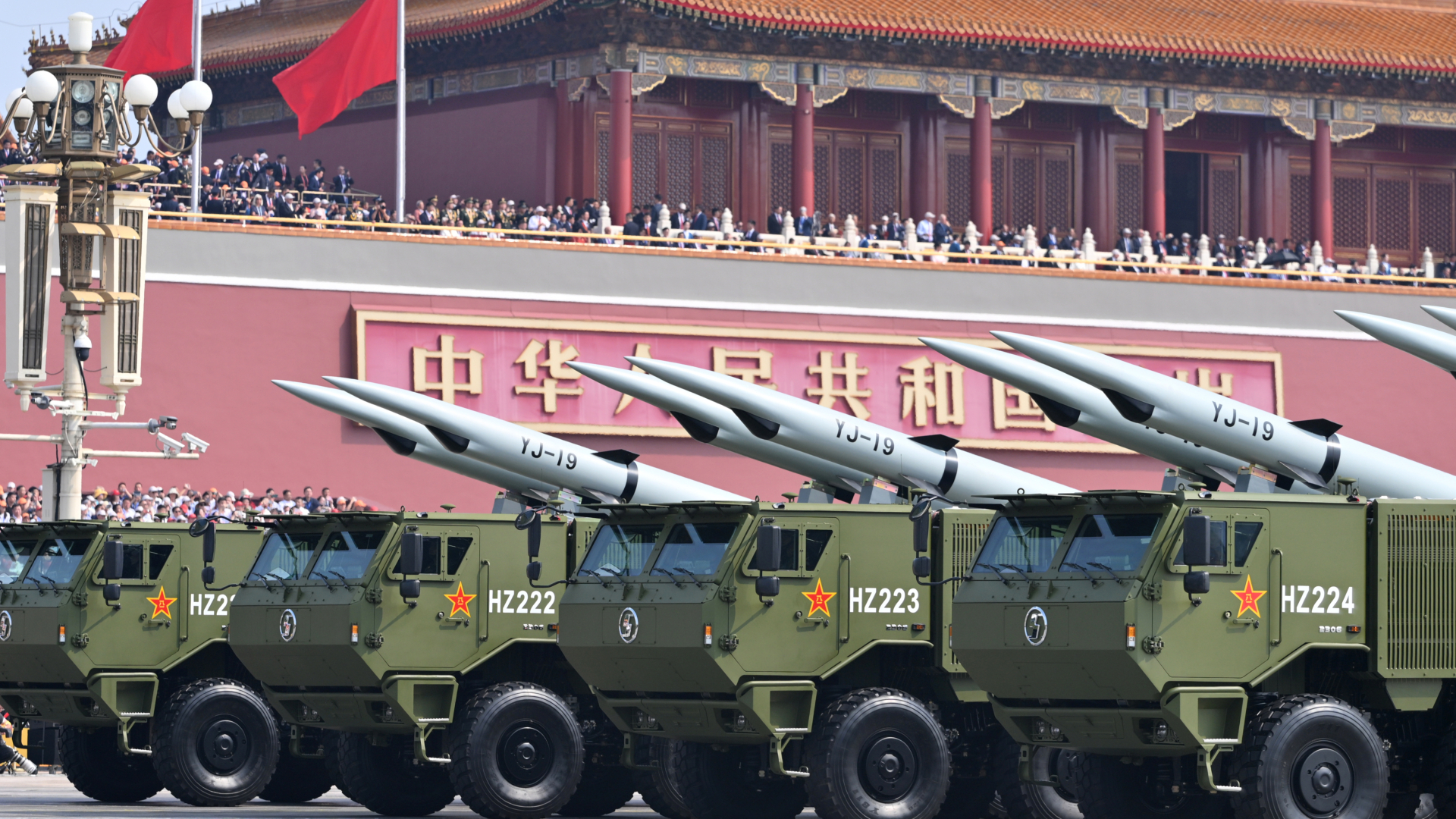 A series of green tanks holding large missiles roll on the streets below a large red building where people are standing next to China&#039;s national flags. 