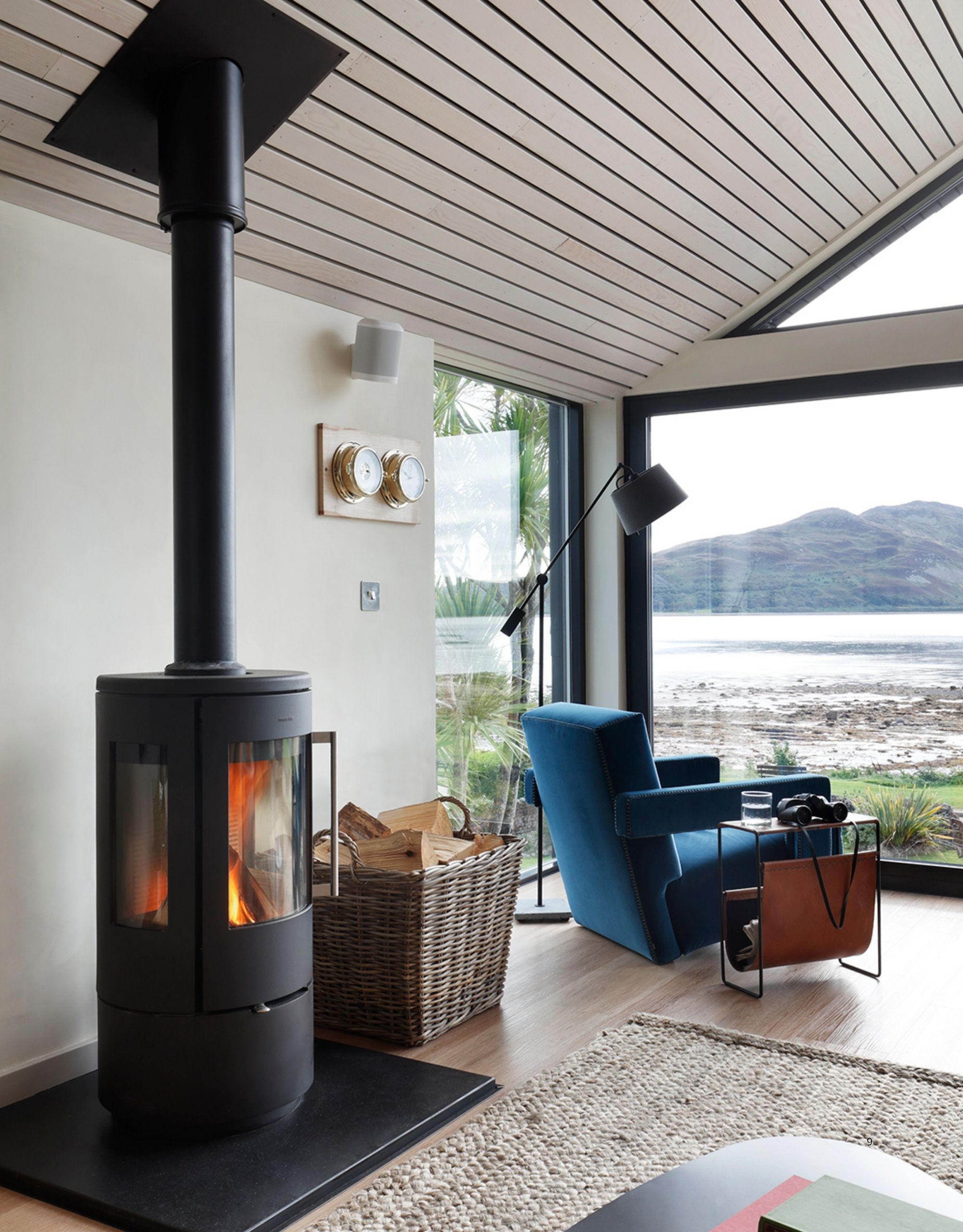 A minimalist, white living room with a large window looking out to a mountainous lake view. There is a fireplace, a basket of wood, a blue chair, a magazine rack, and a lamp by the window.