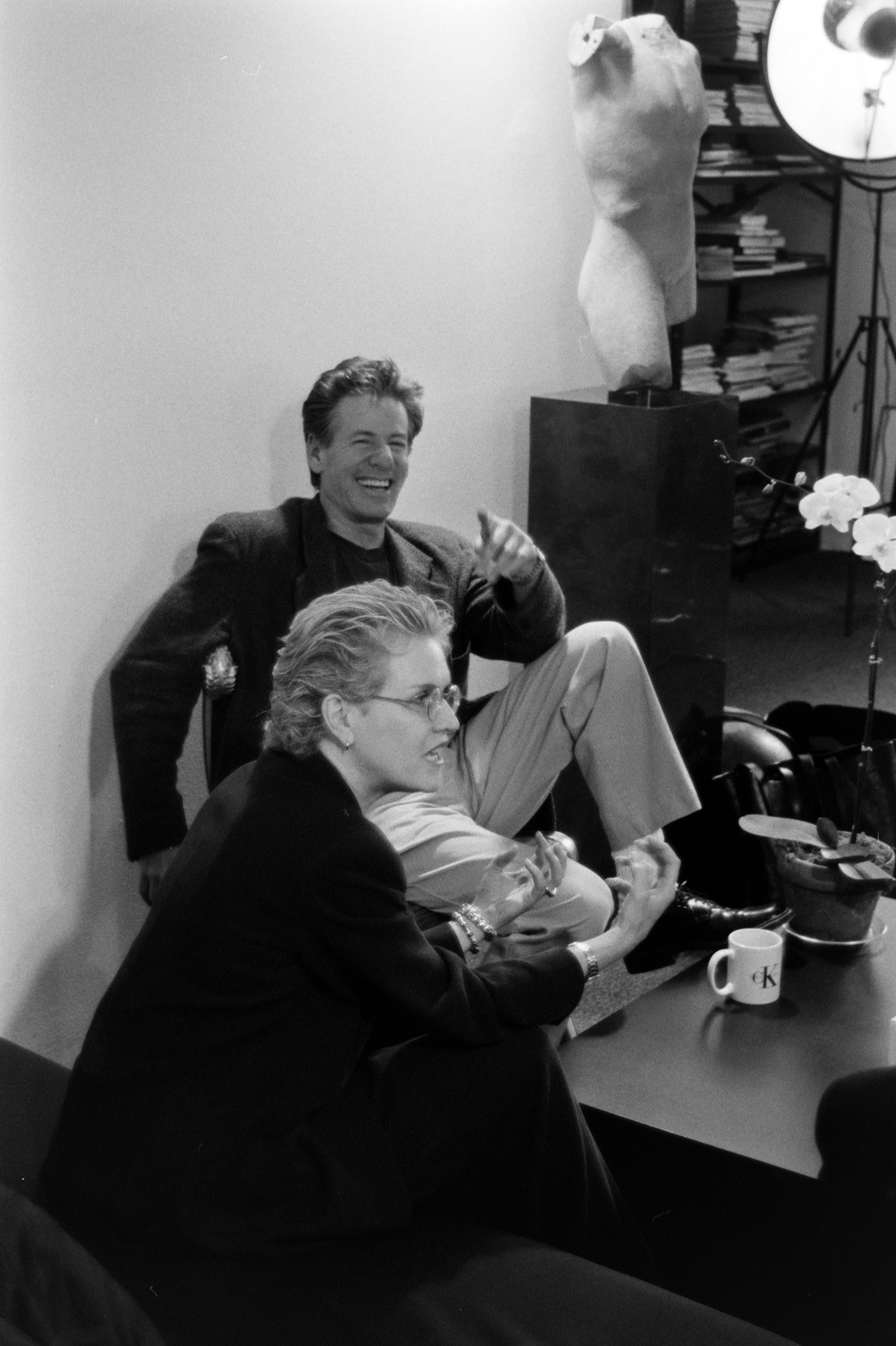 A middle-aged man dressed in a dark tailored suit and beige trousers and a woman dressed in a full black tailored suit talk and laugh in an office decorated with white potted orchids, books, and marble busts.