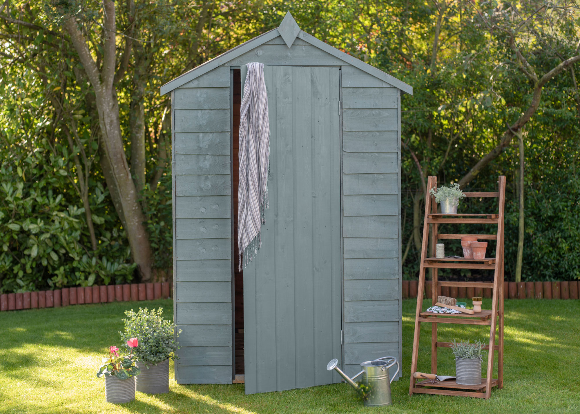 small garden shed painted green with planters, watering can and ladder shelf on lawn area with trees in background