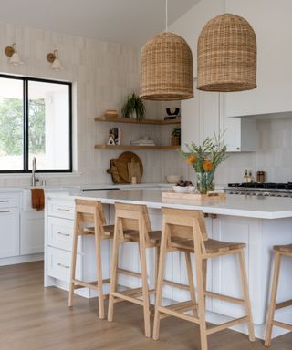 A kitchen with white cabinets, cream wallpaper, wall sconces on the wall, a black window, and a white kitchen island with a wooden tray of flowers on it with four wooden stools underneath it