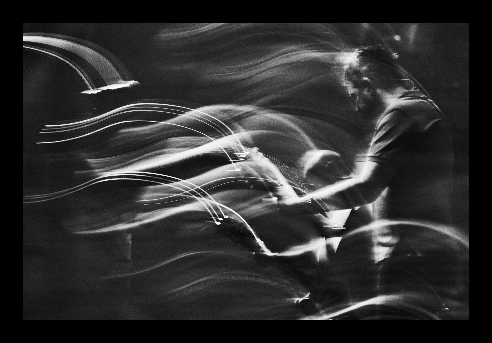 A black-and-white, long-exposure photograph captures a musician in profile, surrounded by dramatic, sweeping light trails that convey high-energy motion.