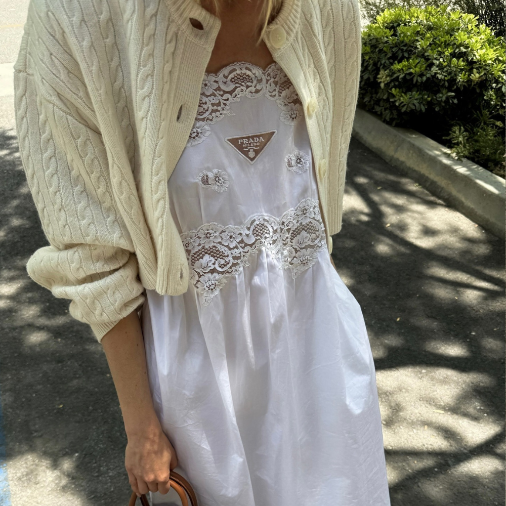 Elsa Hosk stand outside in a park wearing a white cotton lace dress from Prada, which she layered with a butter yellow cardigan and styled with a Hermes bag and animal print shoes.