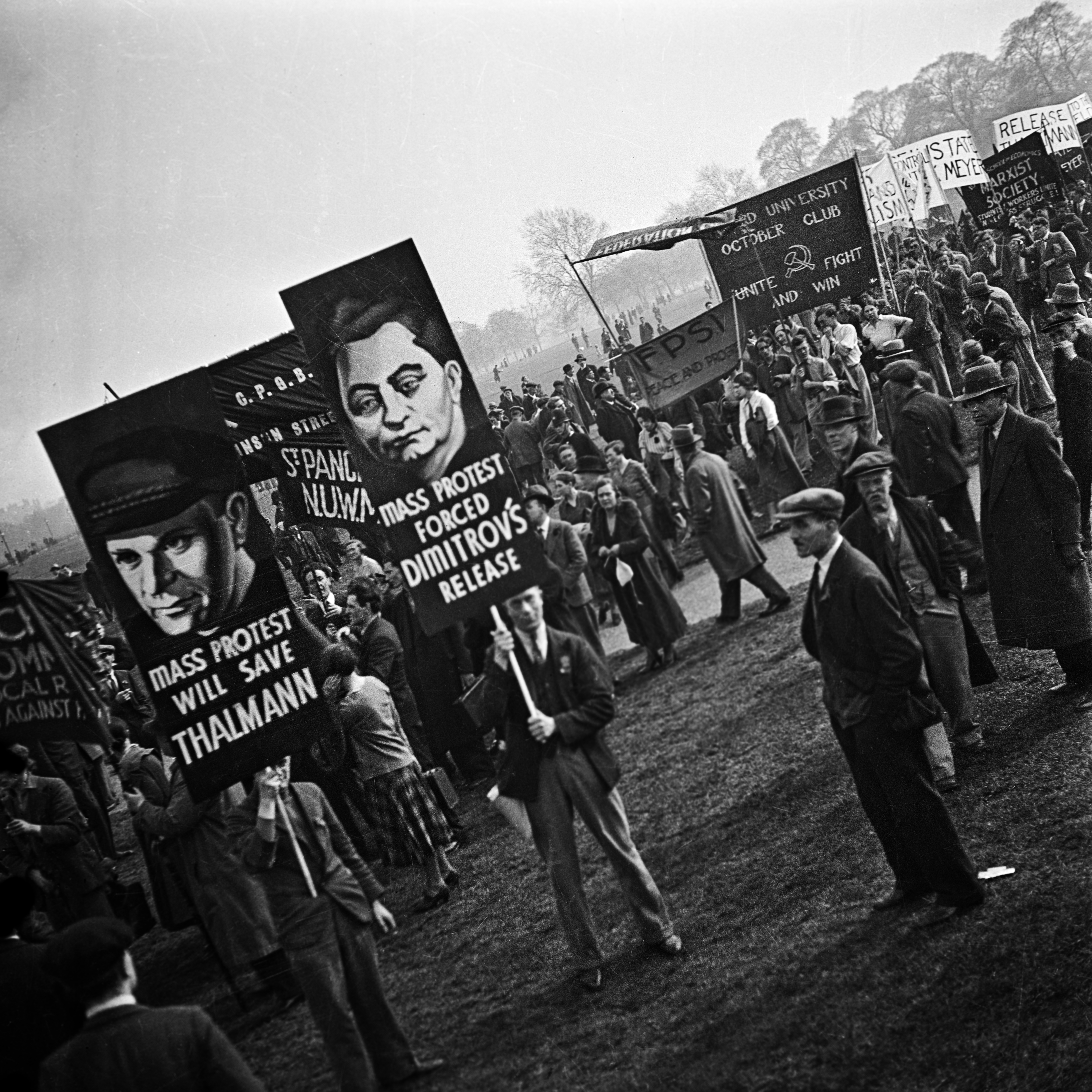 A large crowd marches across open parkland carrying placards bearing painted portraits and slogans including &amp;quot;Mass protest will save Thalmann&amp;quot; and &amp;quot;Mass protest forced Dimitrov's release&amp;quot;. Banners further back reference the Oxford University October Club and a Marxist Society. The scene is overcast, the crowd dense and purposeful.