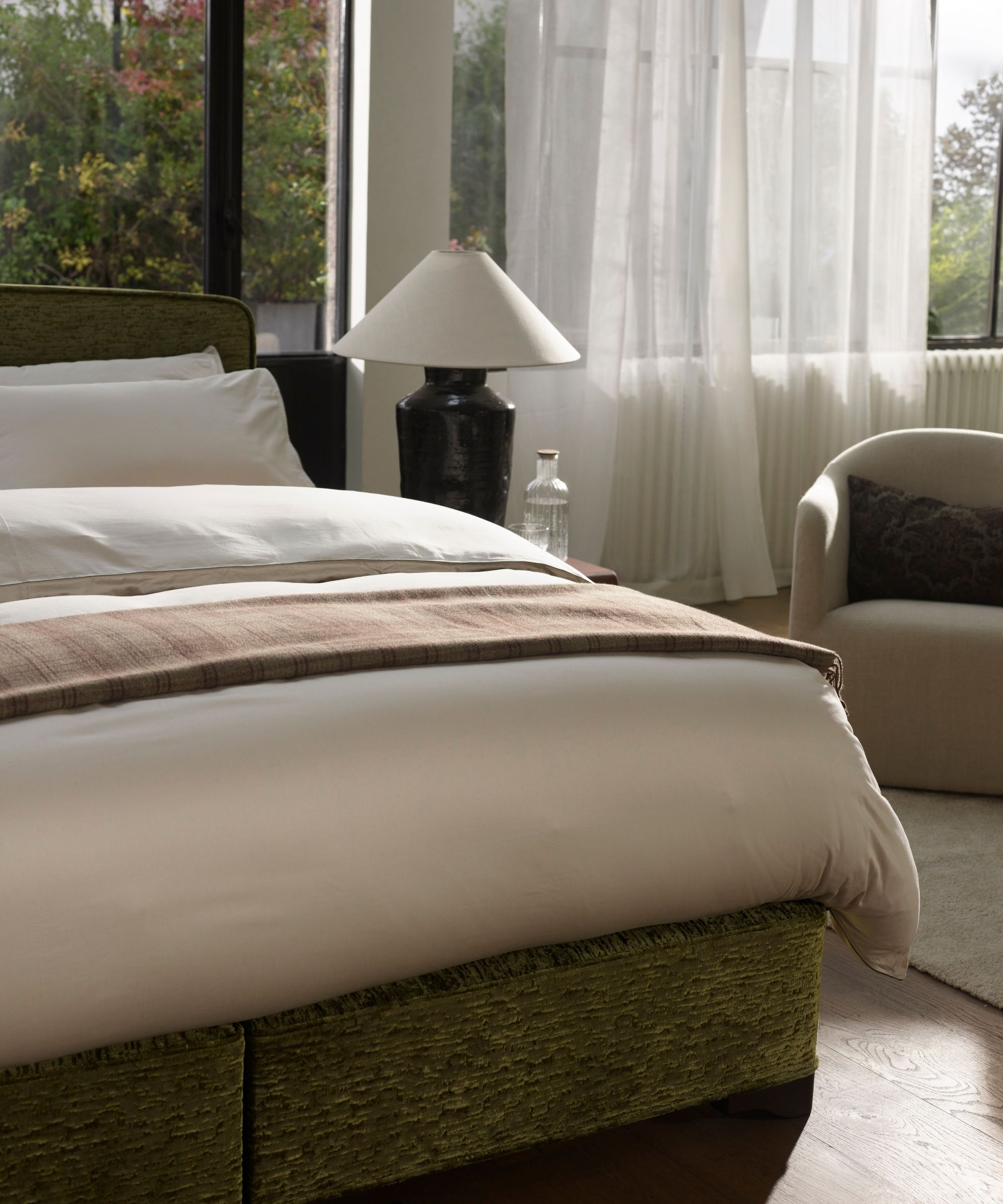 A bright bedroom with large windows with sheer white curtains, a green bed, white bedding, a black lamp with white lampshade, and a large upholstered chair. The flooring is pale brown wood.