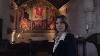 Claire Goose in a dark jacket as Emma Lavenham stands in front of a religious painting in a church.