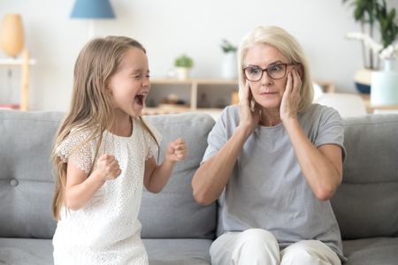 Shocked grandma closing ears not to hear noisy stubborn fussy little granddaughter screaming demanding attention, preschool spoiled kid girl yelling at grandmother, child tantrum manipulation concept