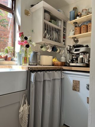 the corner of a kitchen with exposed shelving and a curtain next to a belfast swink to disguise the washing machine