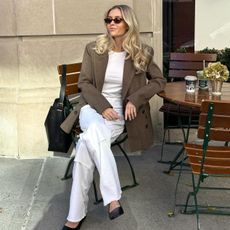 style influencer Fredrika Ekerot sitting in a seat at an outdoor cafe in NYC wearing a brown blazer, white t-shirt, white pants, and black shoulder bag