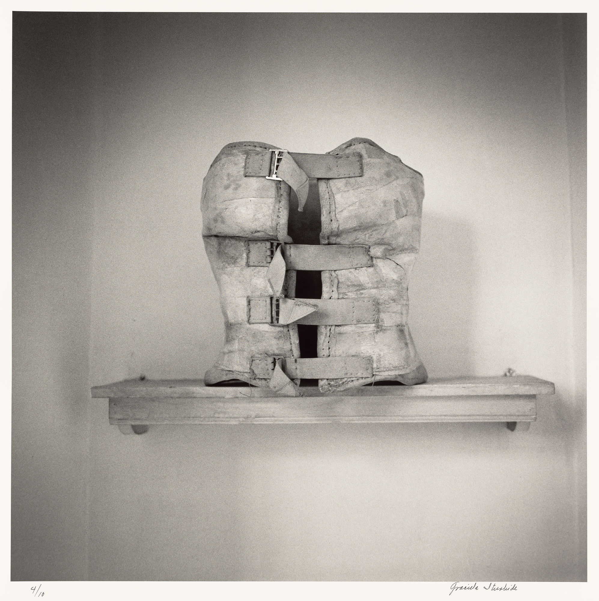 Black and white photograph of a weathered leather back brace with metal buckles, displayed upright on a wooden shelf against a plain wall.